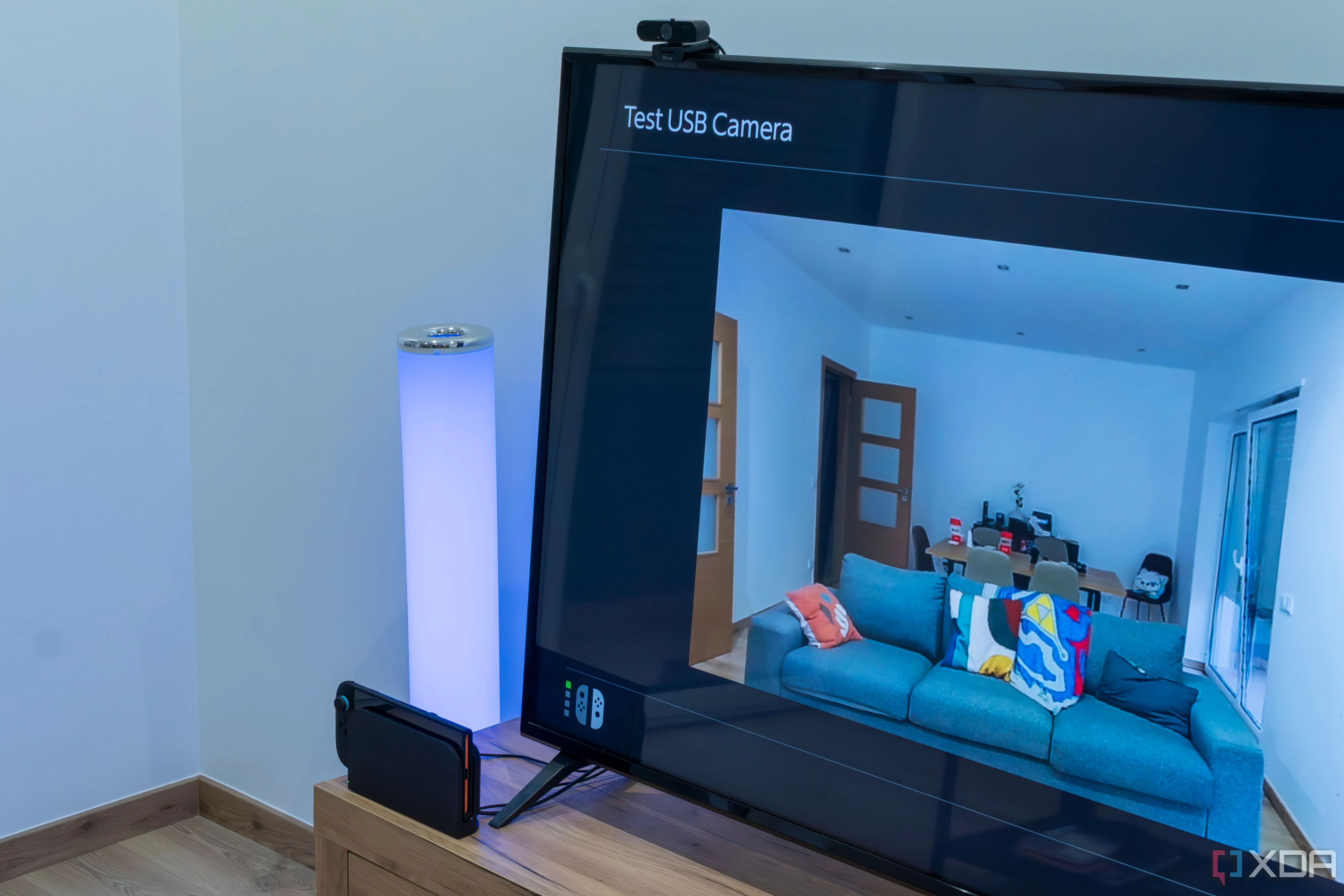Một chiếc Nintendo Switch 2 đặt trong dock, với một webcam Trust USB màu đen cắm vào cổng USB, màn hình TV phía trước hiển thị hình ảnh từ webcam với giao diện GameChat